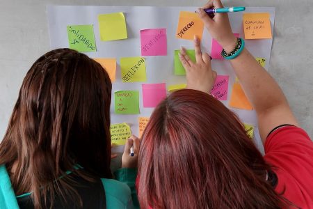 Dos mujeres de espalda escriben papeles pegados a la pared como identidad, solaridad, reflexión.